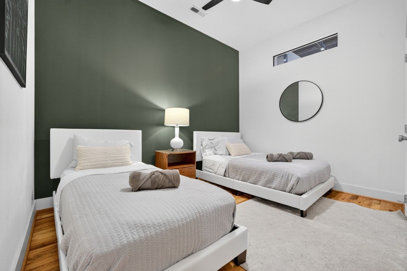 A modern bedroom with two beds, a bedside table with a lamp, and a round mirror on the wall.