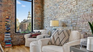 A cozy living room with a brick wall, a stack of books, a white sofa, and a lamp. - Luxury Loft DWTN | Whiskey Trail + River + Lake (Fayetteville)