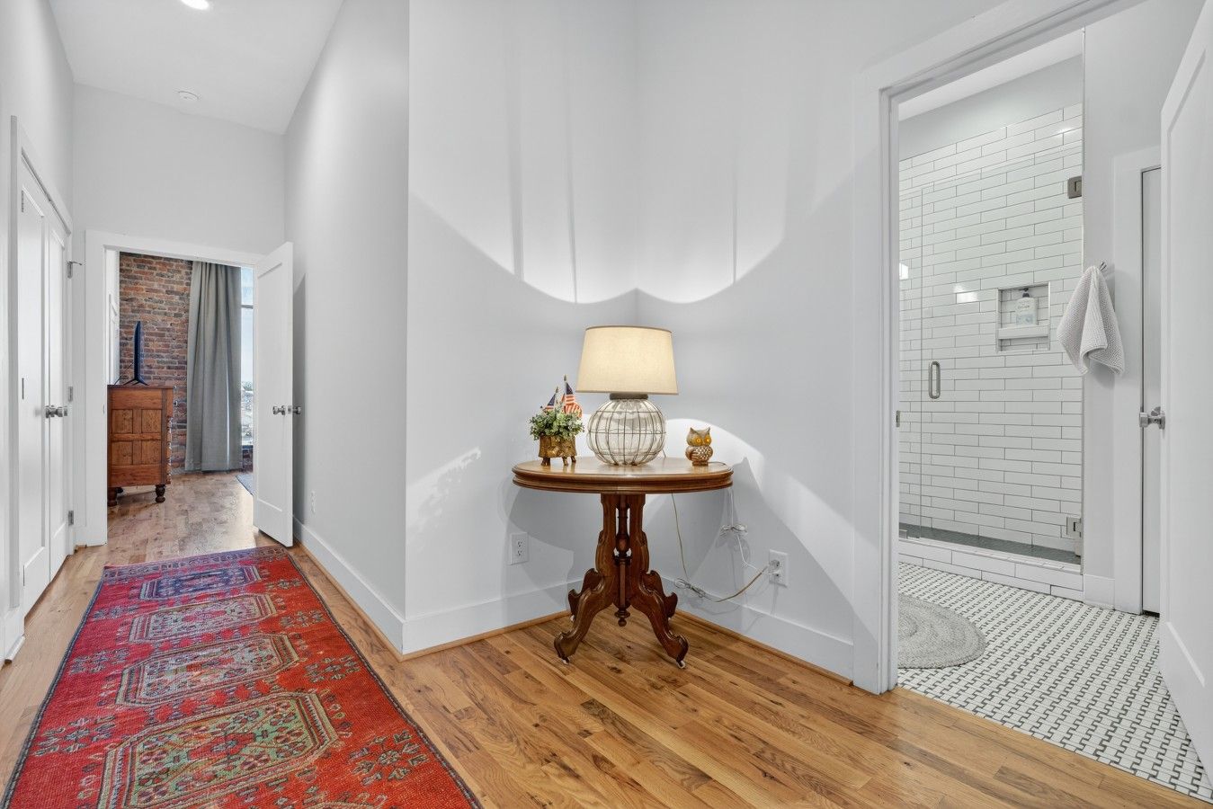 A hallway with a wooden floor, a red patterned rug, a table with a lamp, and a white door leading to a bathroom with a shower.