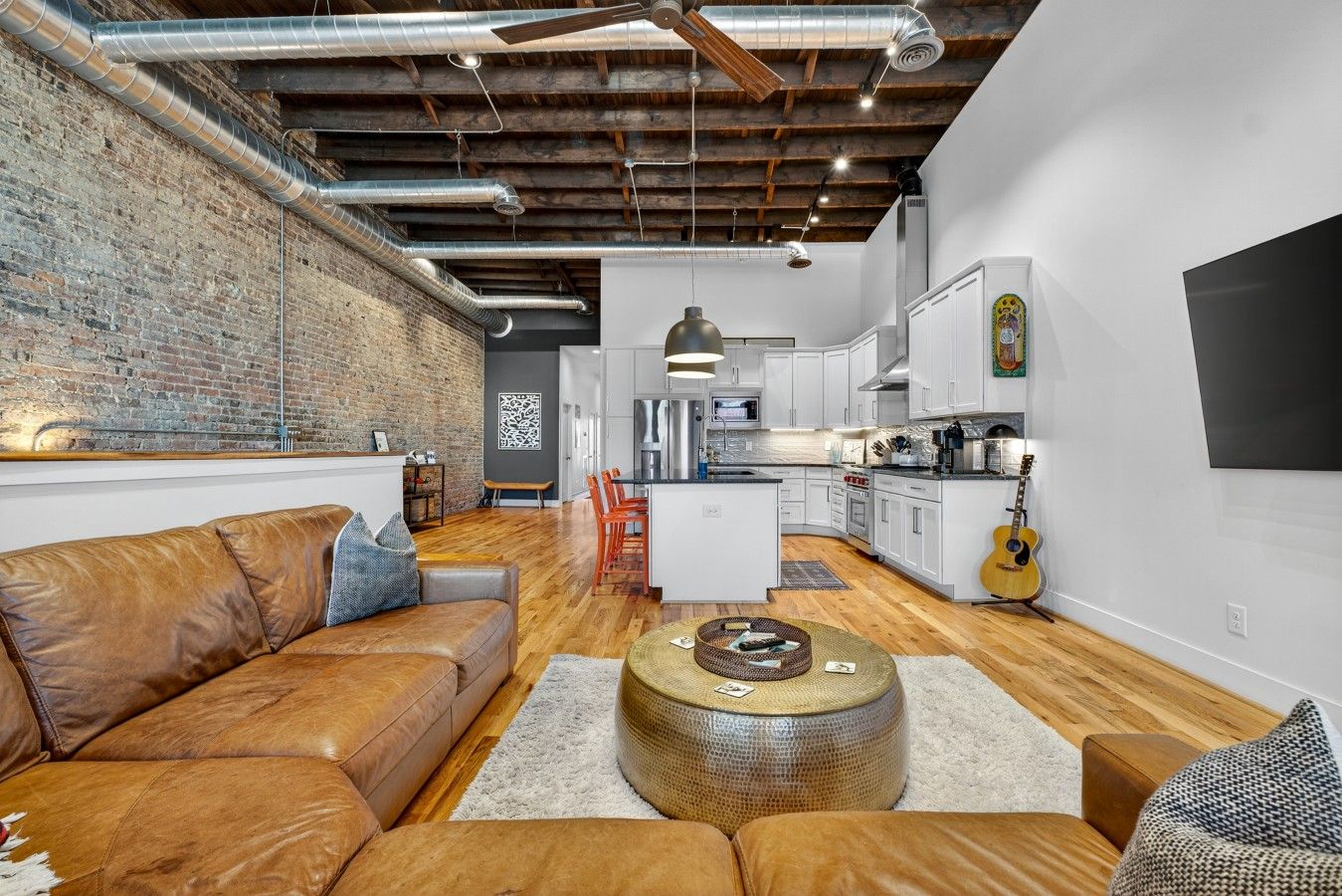 A modern living space with a leather sofa, a round coffee table, and a kitchen area with white cabinets and a guitar.