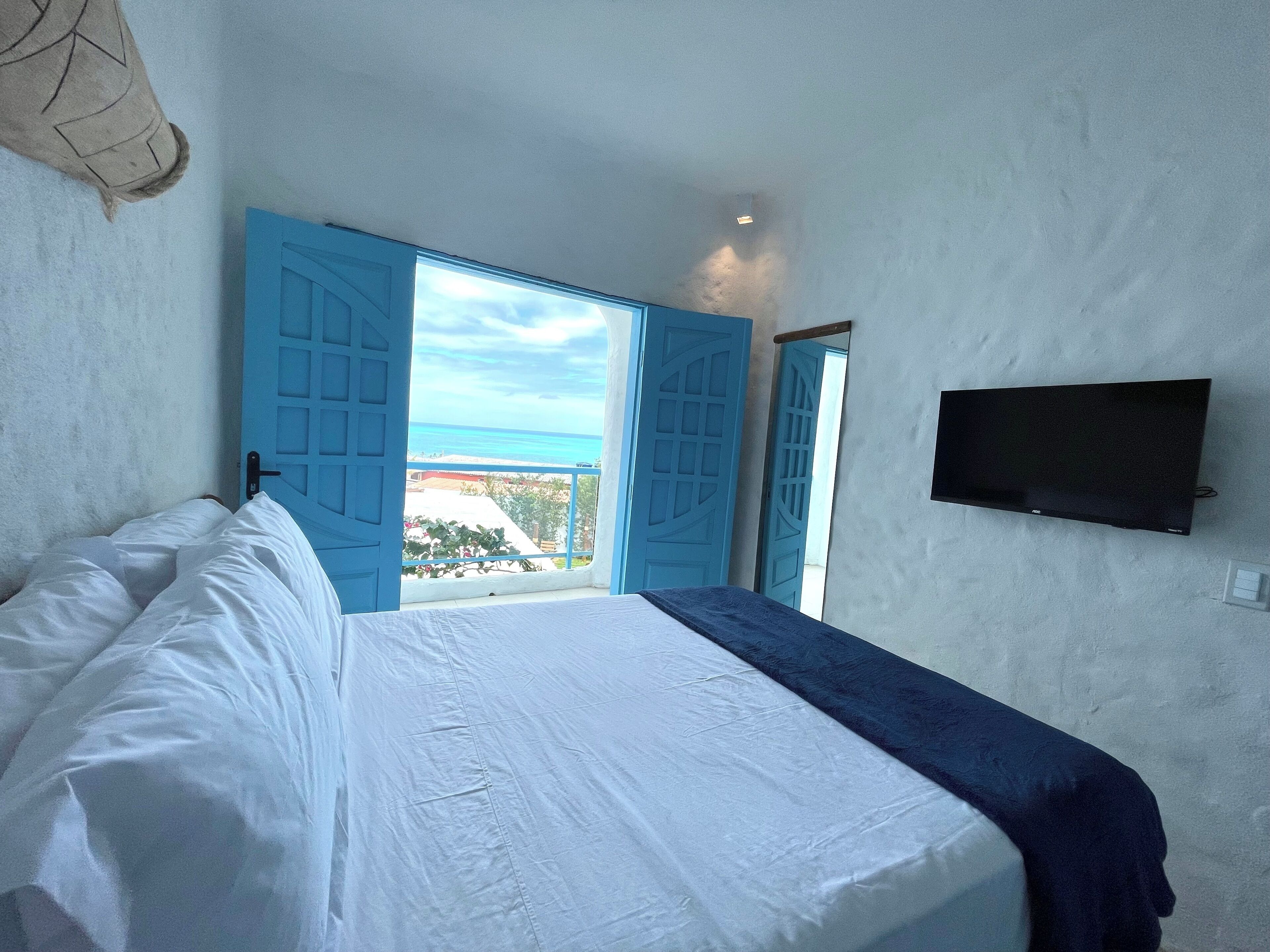 Un dormitorio con cama, televisor montado en la pared y vista al mar a través de puertas azules abiertas.