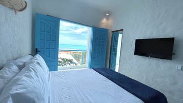 Un dormitorio con cama, televisor montado en la pared y vista al mar a través de puertas azules abiertas.