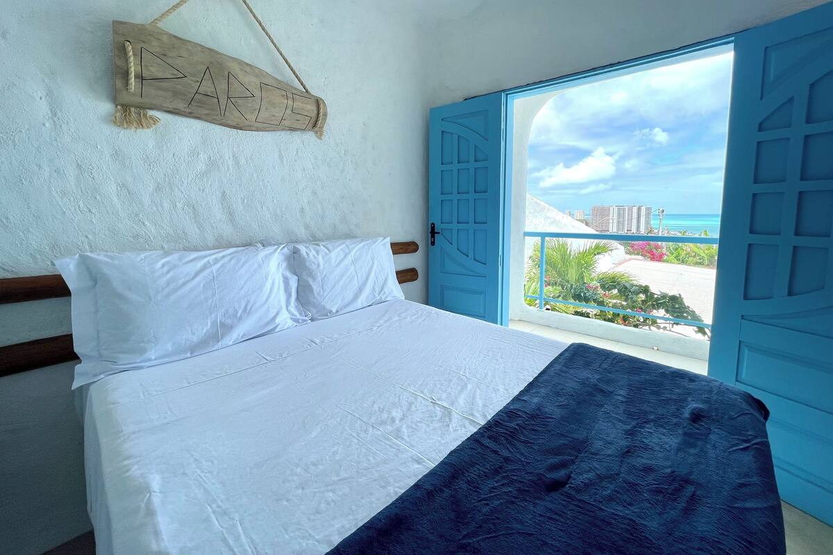 A bed with white and dark blue bedding, a wooden sign with letters, and a view of the sea through open blue doors.