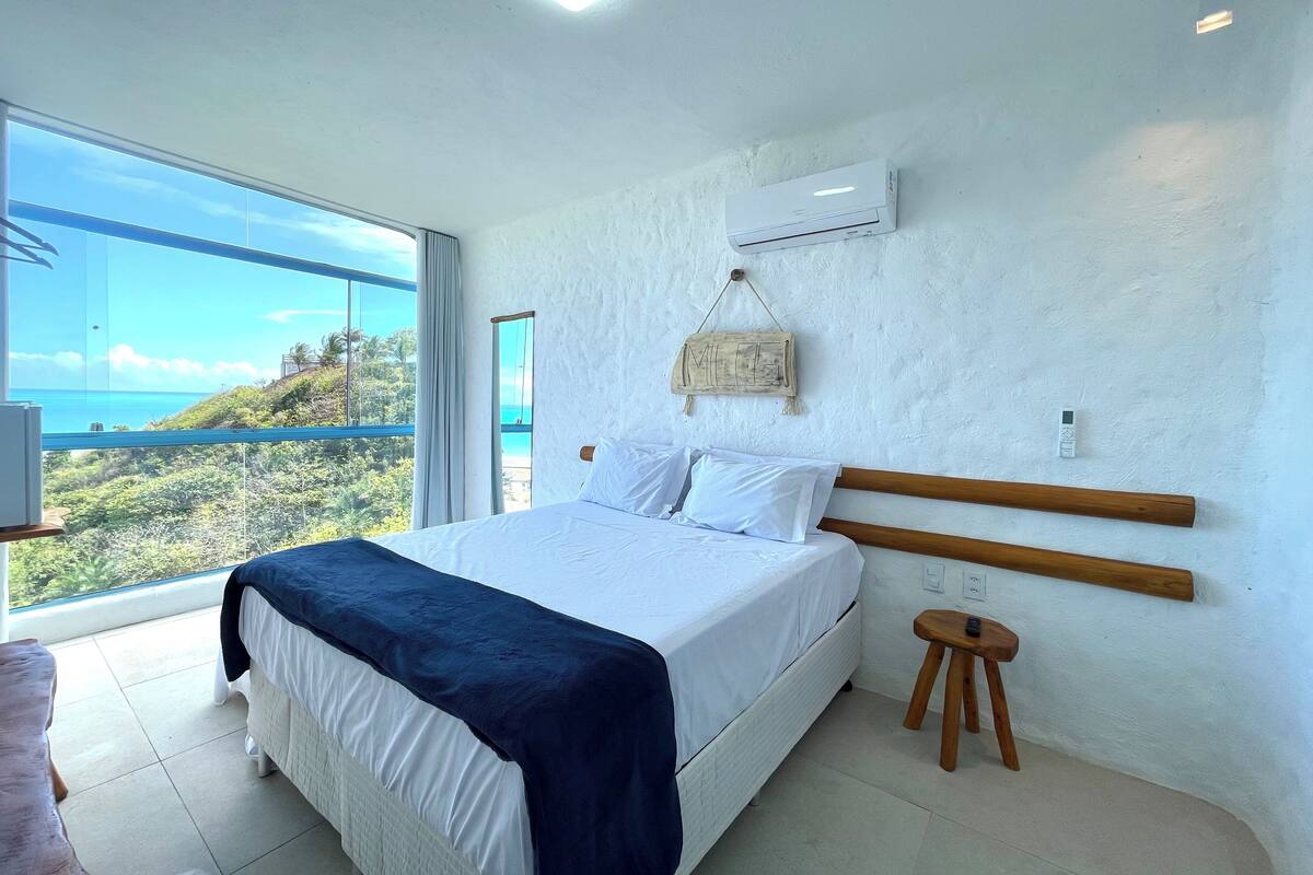 A bedroom with a large bed, a wooden headboard, a small wooden stool, a wall-mounted air conditioner, and a view of the ocean through large windows.