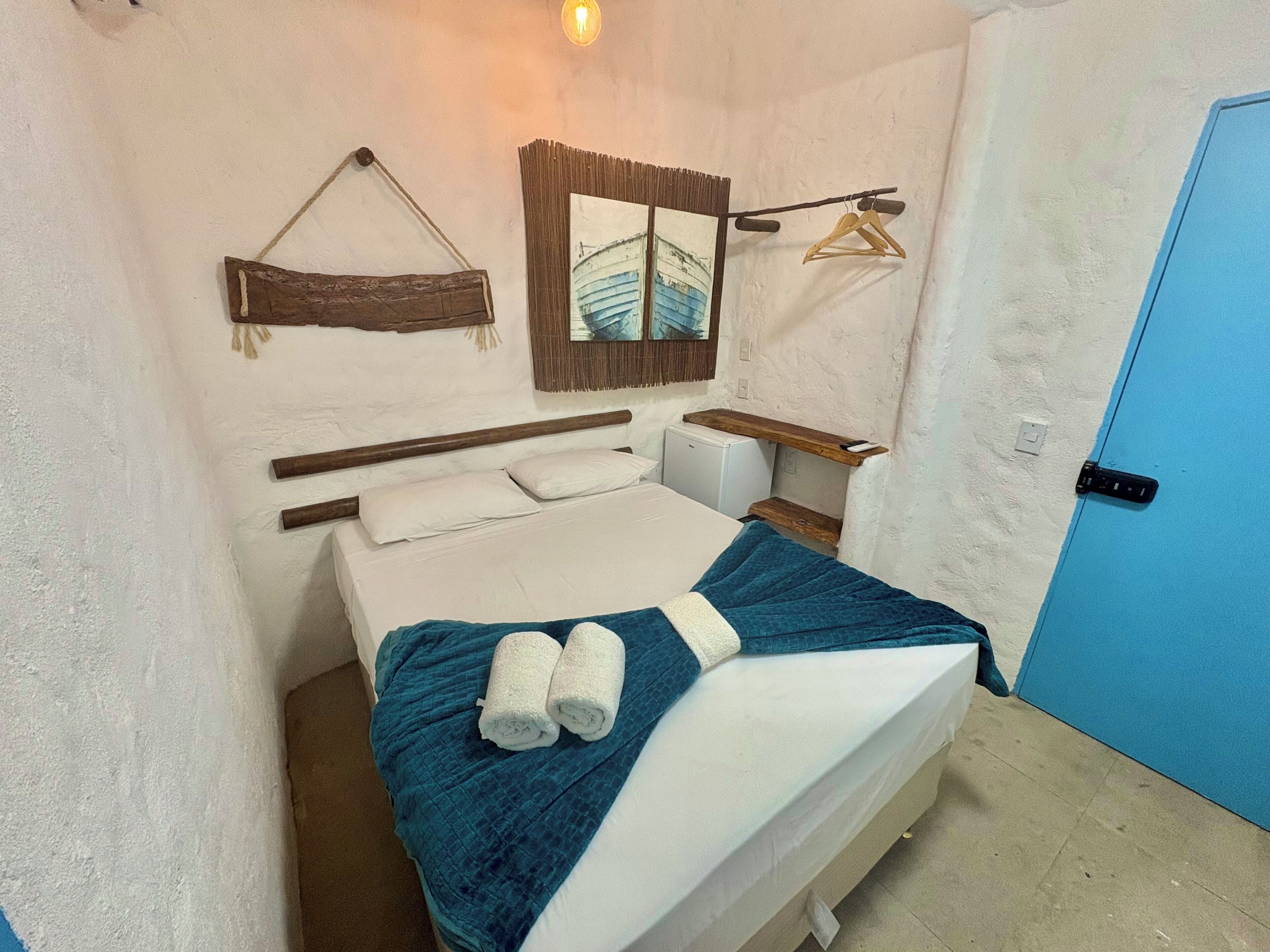Ein Einzelbett mit blauer Decke und zwei zusammengerollten Handtüchern, ein hölzernes Kopfende, ein kleiner Nachttisch, ein an der Wand befestigter Spiegel und eine blaue Tür.