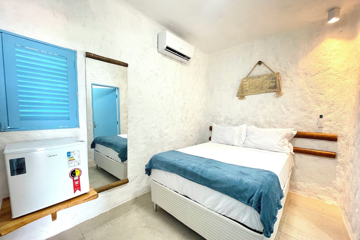 A bedroom with a bed, a white wall, a mirror, an air conditioning unit, and a small shelf with a white appliance.