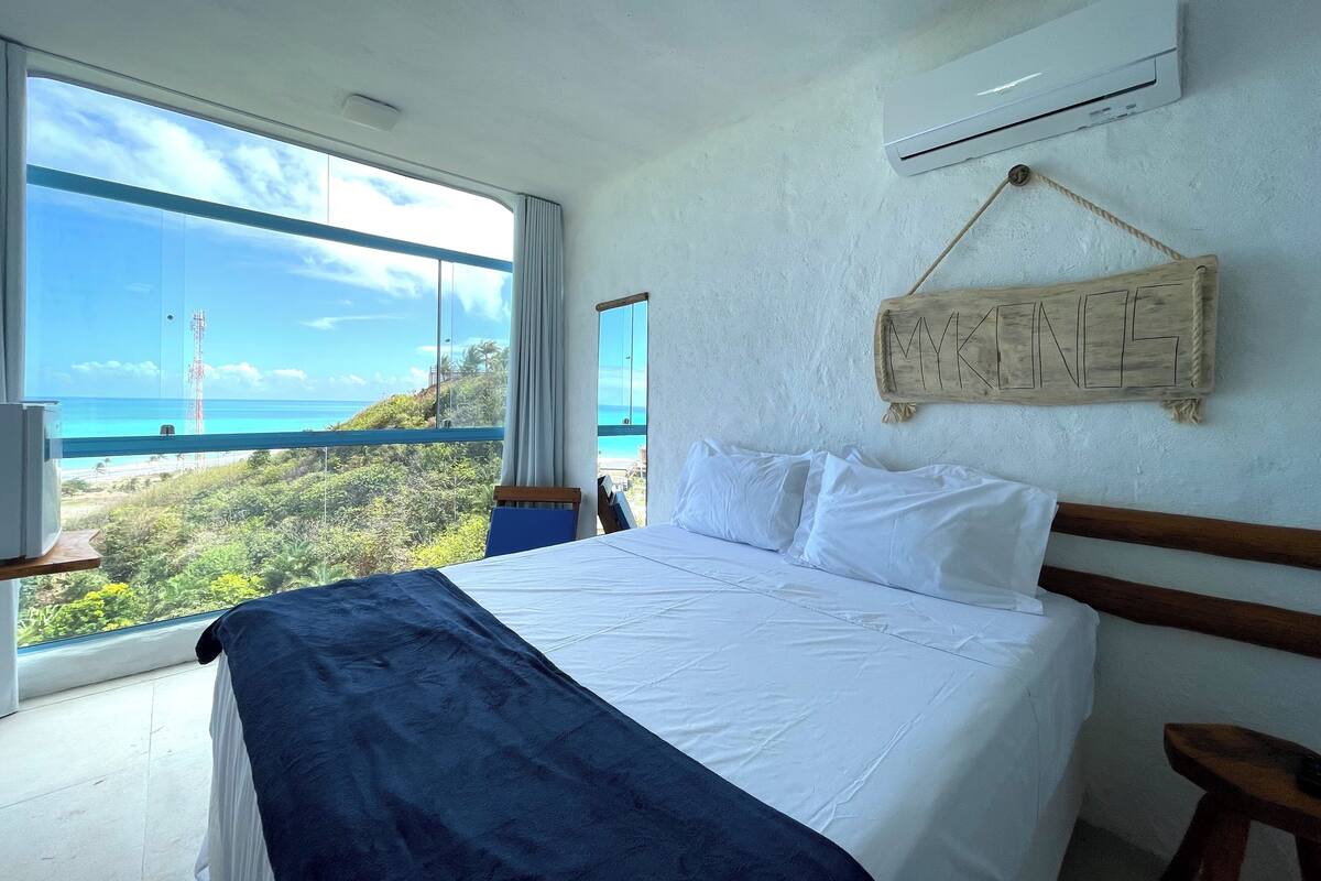 A bedroom with a large window, a bed, a wooden headboard, a wall-mounted air conditioner, and a sign with the word "MYKINGS".