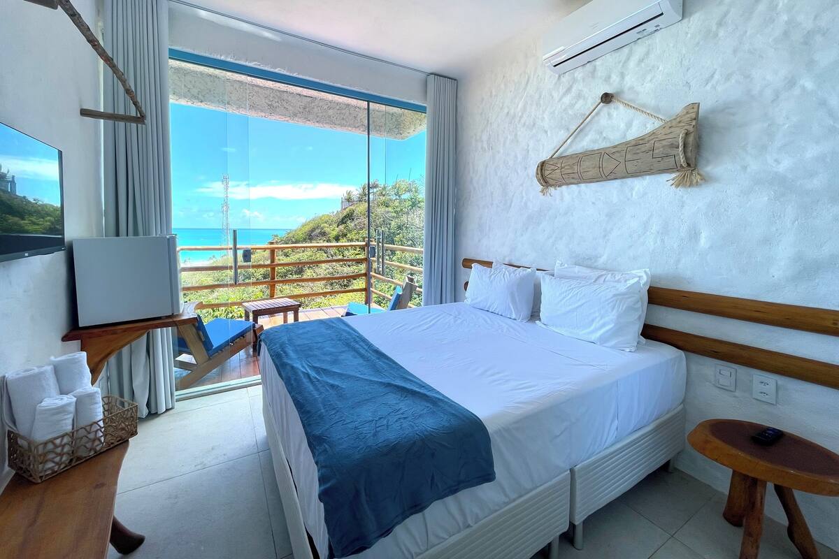 A bedroom with a bed, a bedside table, a small table, a chair, a window with a view of the ocean, and an air conditioning unit.