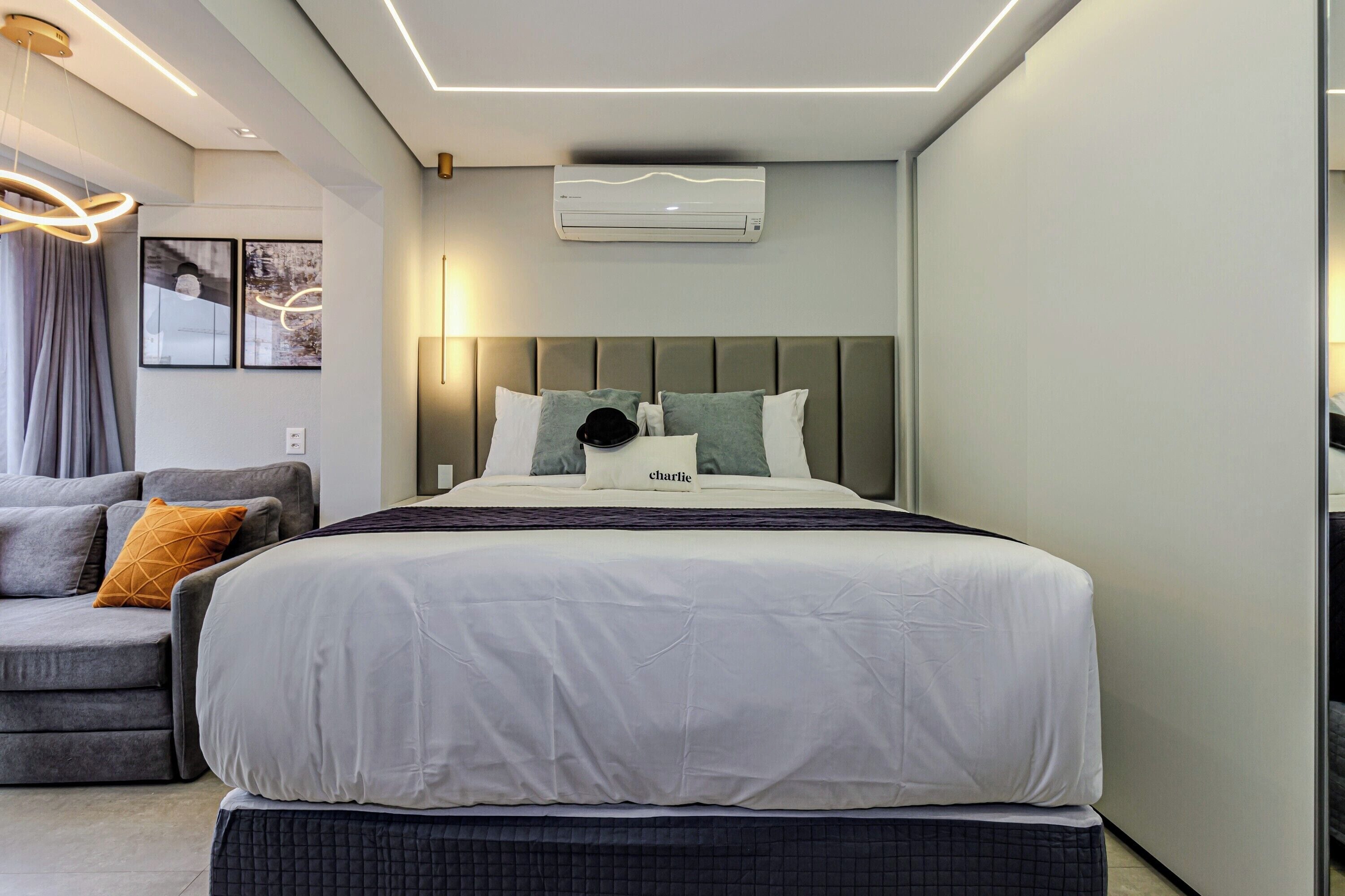 A modern bedroom with a large bed, a grey sofa, and a wall-mounted air conditioner.
