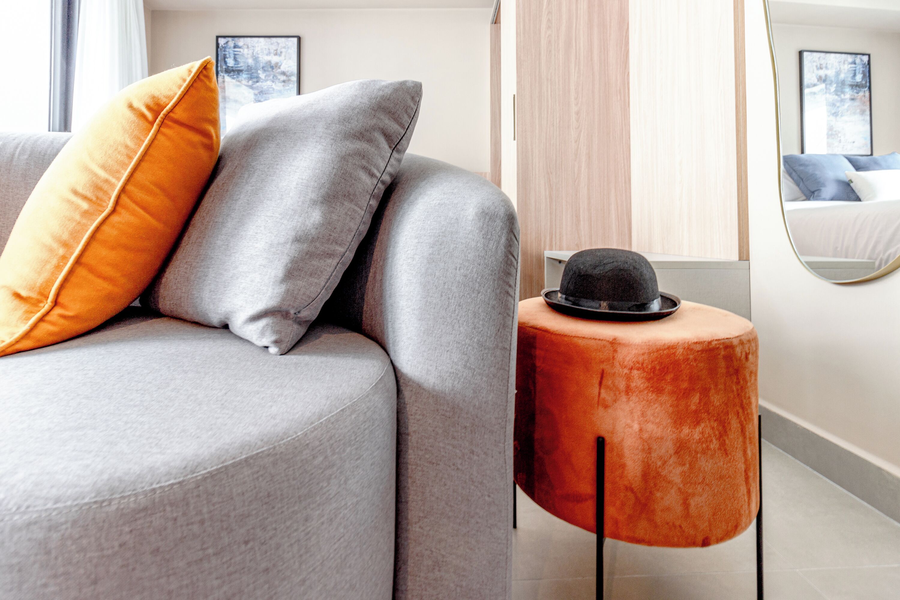 Ein modernes Wohnzimmer mit einem grauen Sofa, orange-grauen Kissen und einem runden, orangefarbenen Beistelltisch mit einem schwarzen Hut.