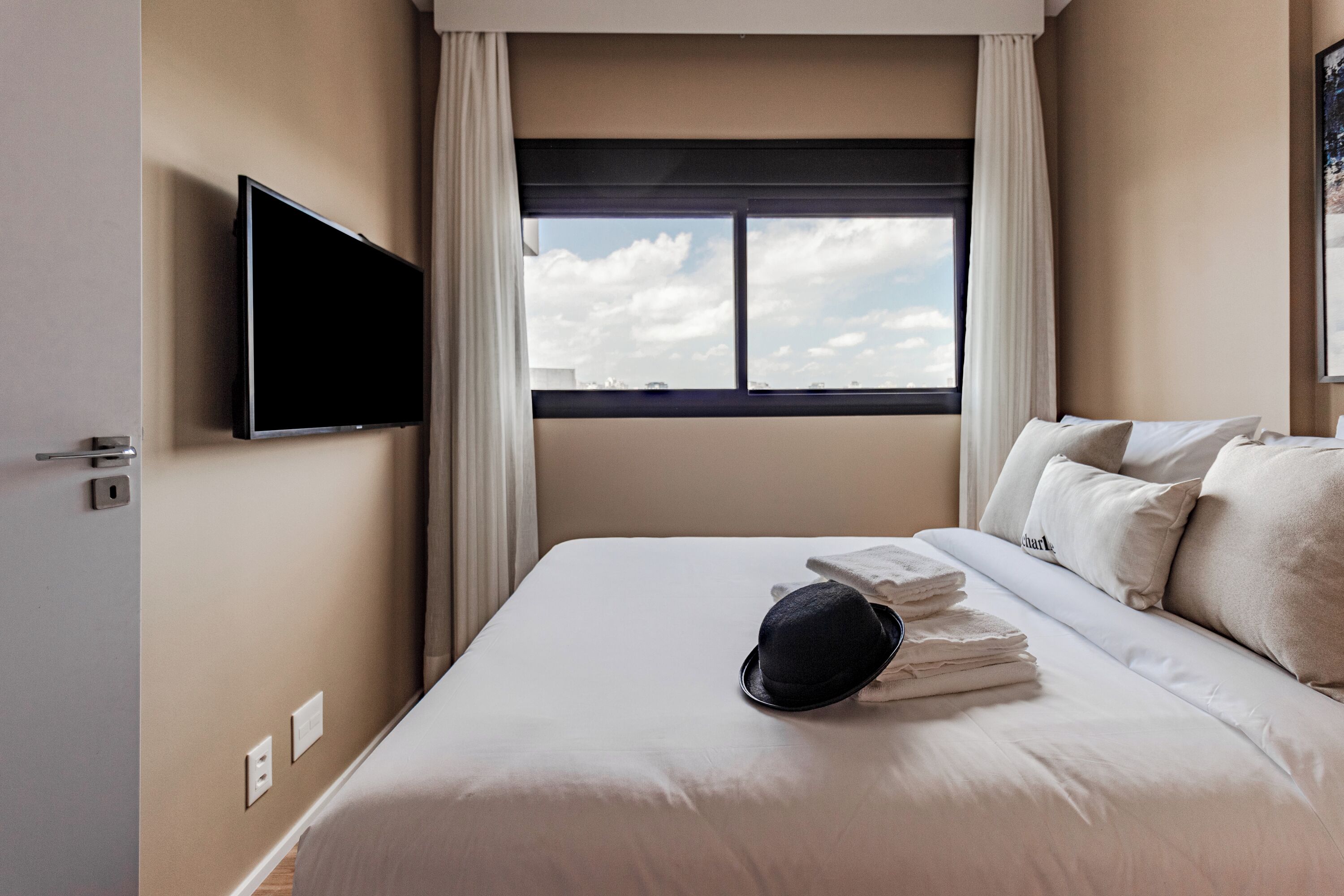 Habitación de hotel con cama, televisor de pantalla plana montado en la pared, ventana con cortinas y vista al cielo.