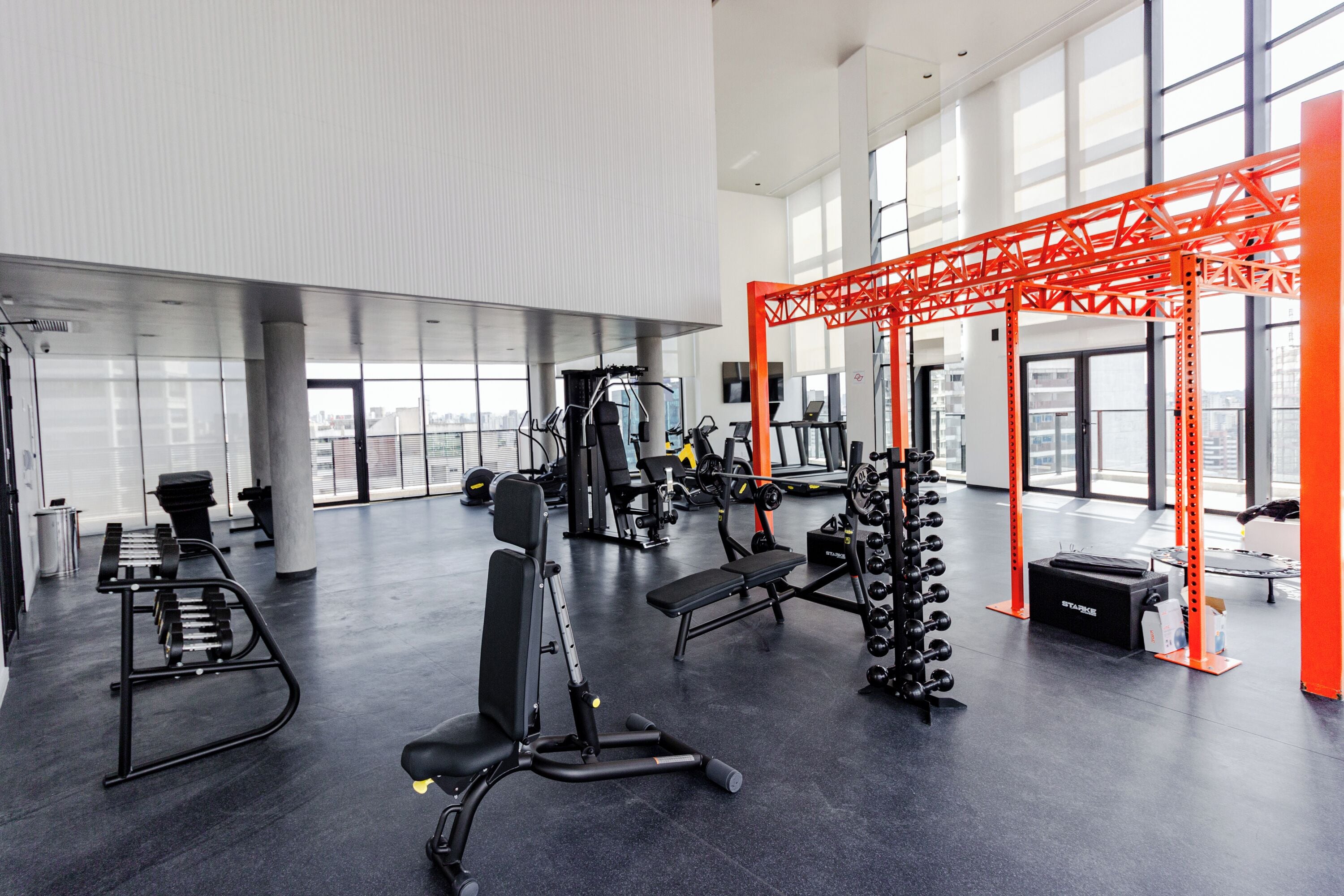 Un gimnasio amplio con diverso equipo de ejercicio, ventanas grandes y un elemento estructural rojo característico.