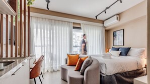 Habitació d'hotel moderna amb un sofà corbat, un llit amb nombrosos coixins, una taula de treball i una finestra gran amb cortines transparents.