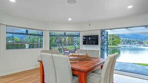 A dining area with a wooden table and six chairs, large windows with a view of a lake and mountains, and a modern kitchen visible through the glass door.