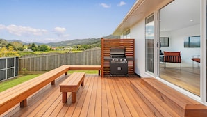 Eine Holzdielen-Terrasse mit Grill, Sitzbank und Glastür zu einem Raum mit Fernseher und Sitzgelegenheiten.
