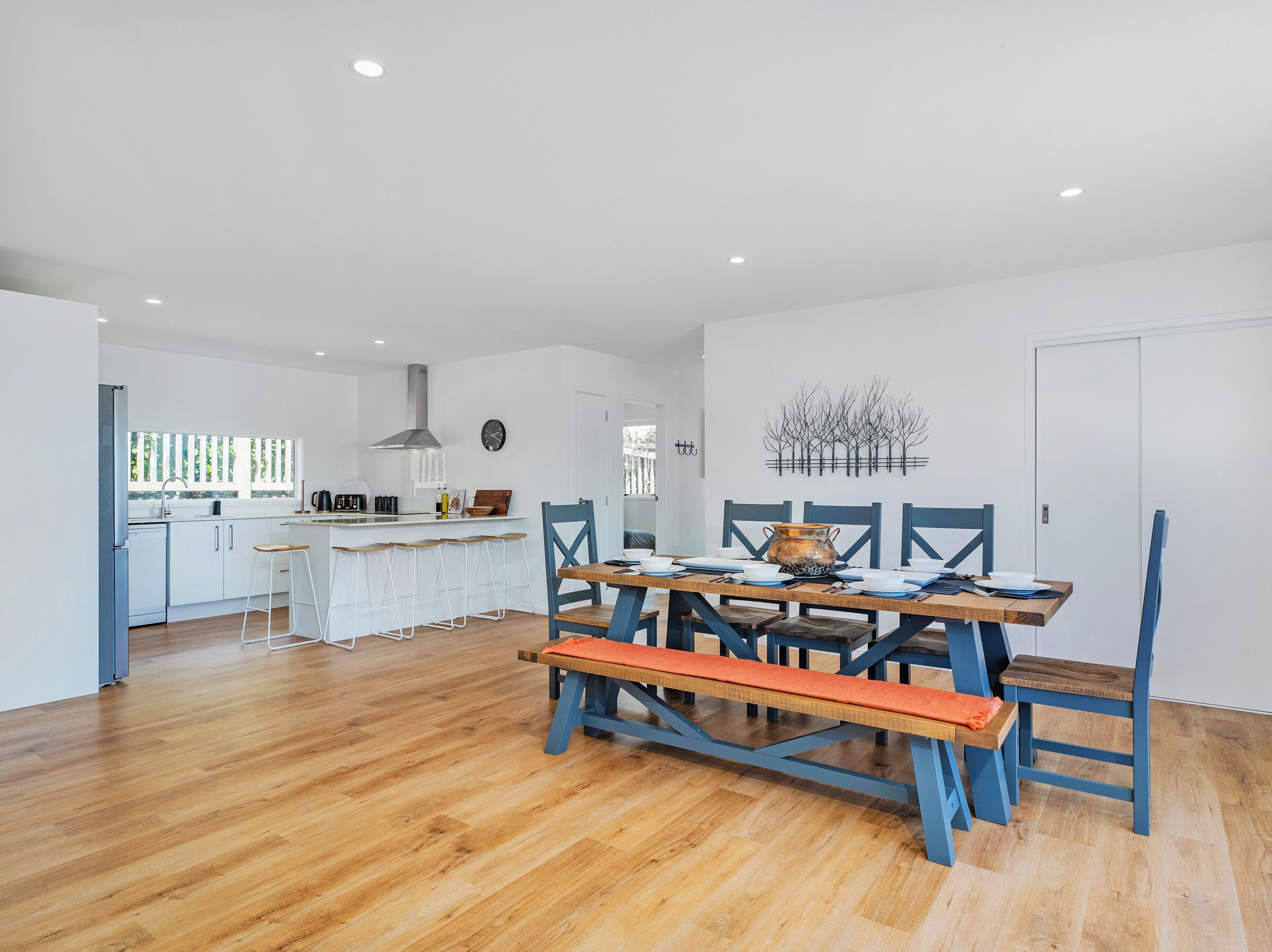 Une cuisine moderne avec des meubles blancs, un îlot central et un coin repas comprenant une table en bois et des chaises bleues.