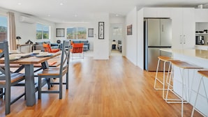 Eine moderne Küche mit einer großen Kochinsel, ein Essbereich mit Holztischen und -stühlen sowie ein Wohnzimmer mit Sofa und Blick ins Freie.