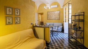 Habitación de hotel compacta con sofá amarillo, zona de comedor con mesa y sillas, candelabro y suelo a cuadros blanco y negro.