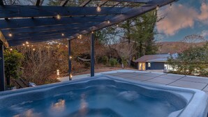 A hot tub under a wooden pergola with string lights, surrounded by trees and a house in the background. - HotTub, FirePit, Pets, Pool Table, Outdoor Dining (Hendersonville)