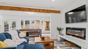 A modern living room with a blue sofa, a wooden coffee table, and a large flat-screen TV mounted on a white brick wall. - HotTub, FirePit, Pets, Pool Table, Outdoor Dining (Hendersonville)
