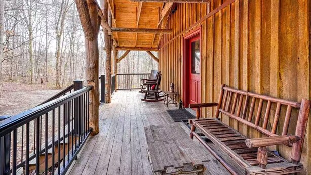 Une véranda en bois avec un banc et des fauteuils à bascule, une porte rouge et une vue sur la forêt environnante.