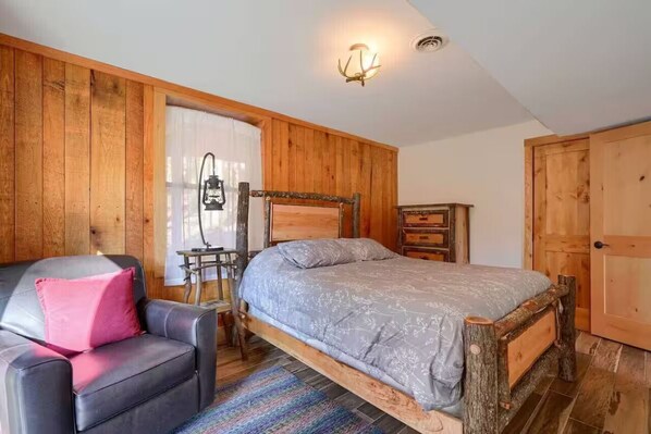 A bedroom with a wooden bed frame, a black leather chair with a pink pillow, a small wooden desk, and a door.