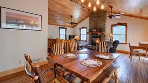 Un coin repas avec une table en bois dressée pour quatre personnes, entourée de chaises en bois. On aperçoit une cheminée en pierre au fond, ainsi que des ventilateurs de plafond et des poutres apparentes.