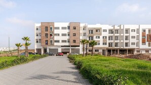 A modern multi-story residential building with a paved driveway, parked cars, and a grassy area with palm trees.