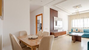 A modern living room with a wooden dining table, beige chairs, a blue sofa, and a TV mounted on the wall.