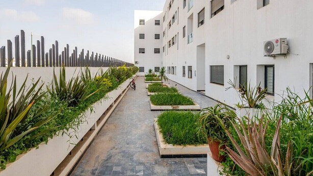 A modern building with a clean, white facade, a narrow pathway lined with planters, and an air conditioning unit on the exterior wall.