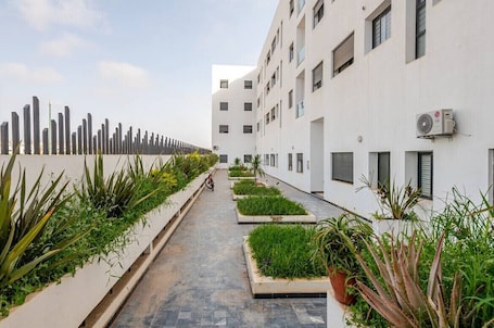 A modern building with a clean, white facade, a narrow pathway lined with planters, and an air conditioning unit on the exterior wall.