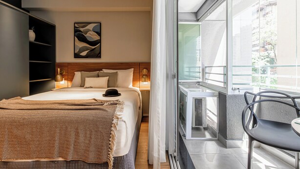 A bedroom with a bed, bedside tables, a wardrobe, a painting, and a balcony with a chair. - Charlie Mode Perdizes (São Paulo)