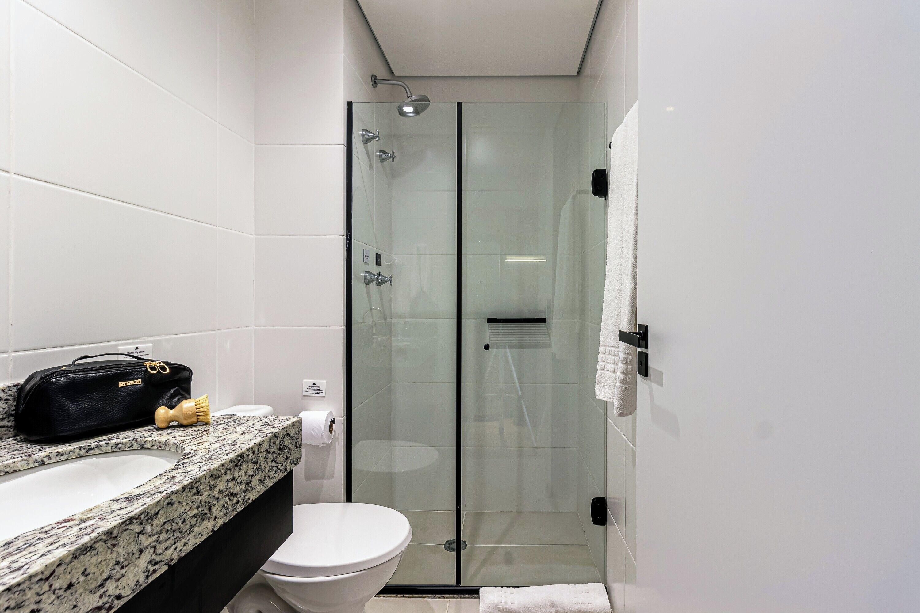 Un baño moderno con cabina de ducha de vidrio, inodoro blanco y encimera de granito con un bolso negro y un cepillo de dientes.