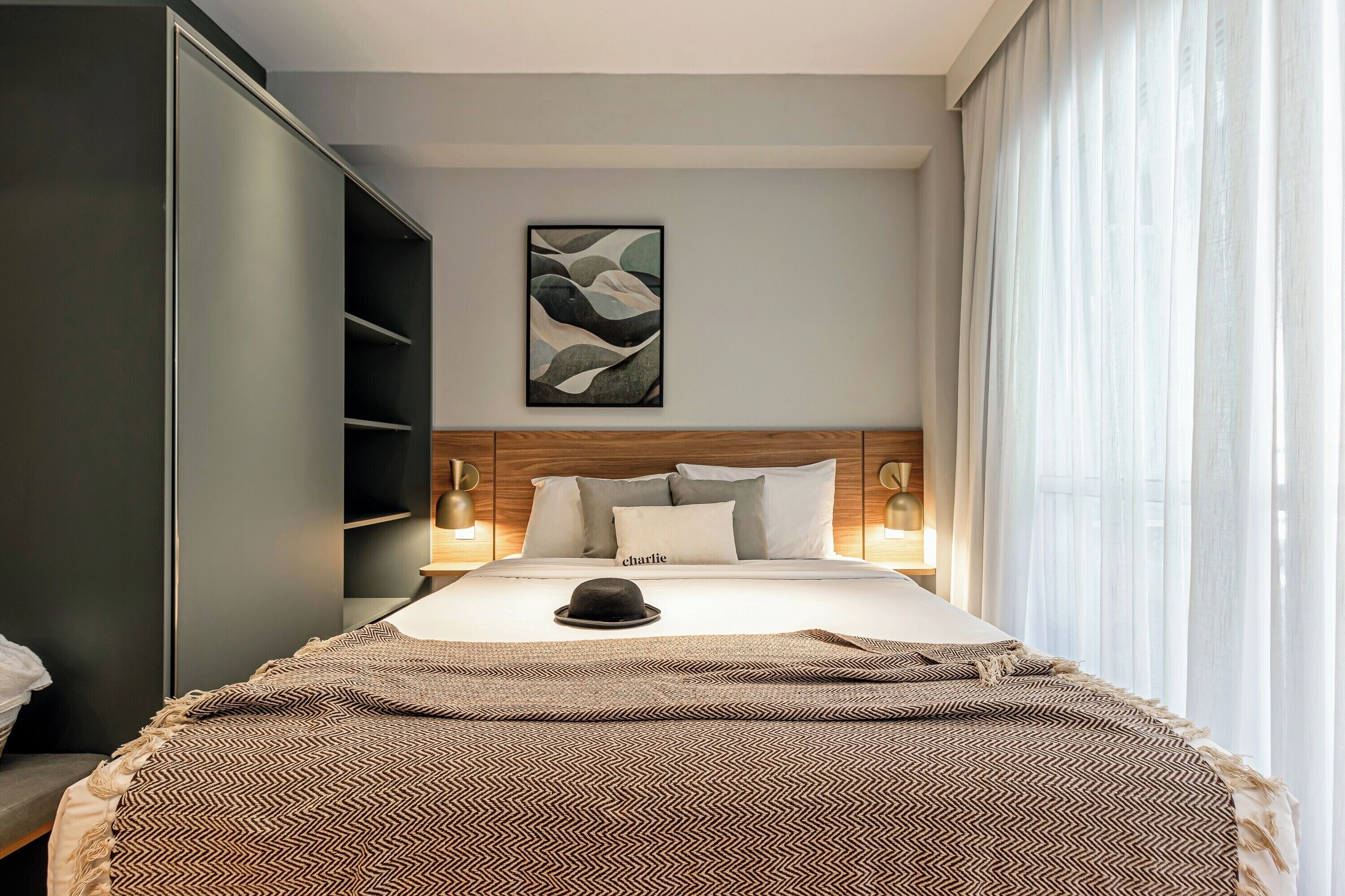 Un letto rifatto con cura, coperto da una coperta a motivi decorativi, un cappello sopra e un quadro astratto appeso alla parete.