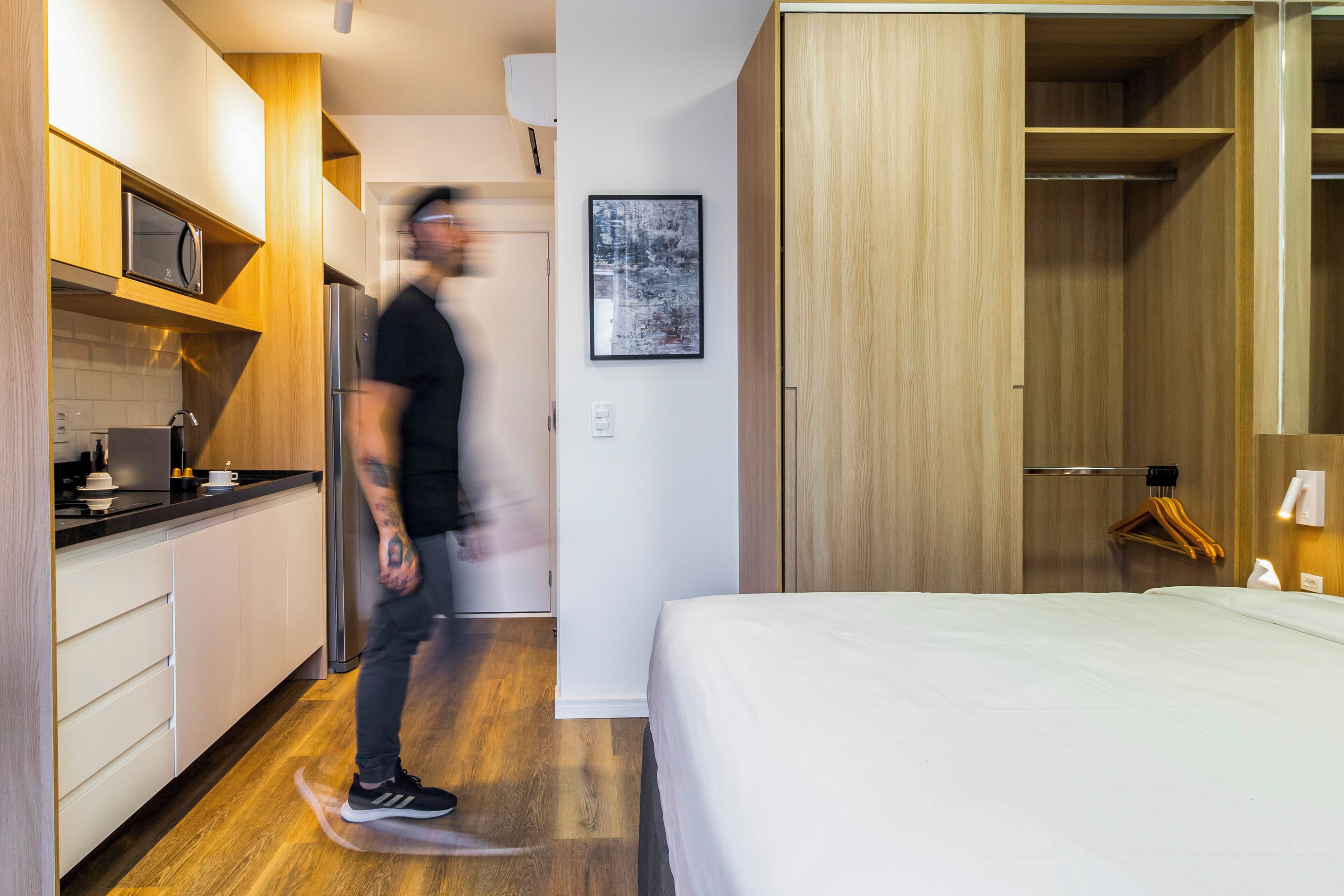 A modern hotel room with a wooden floor, a bed, a kitchenette, and a wardrobe.