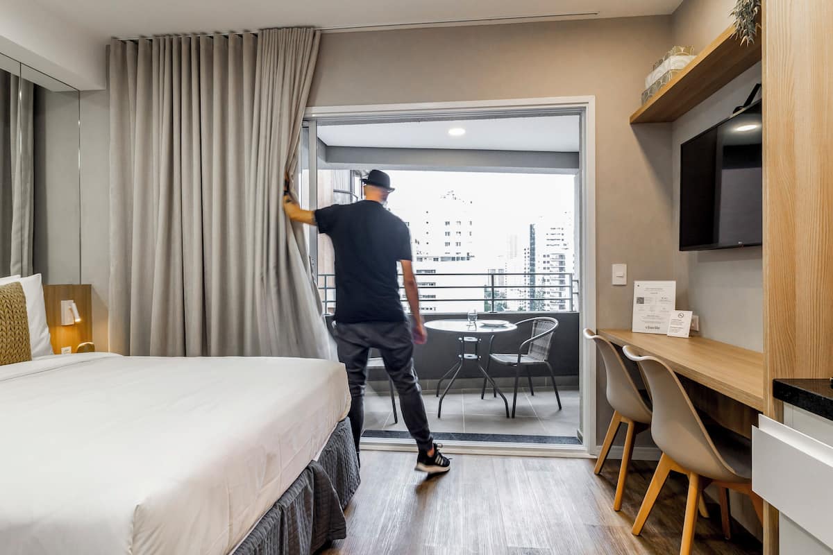 A hotel room with a bed, a desk, a chair, a TV, and a balcony with a city view.