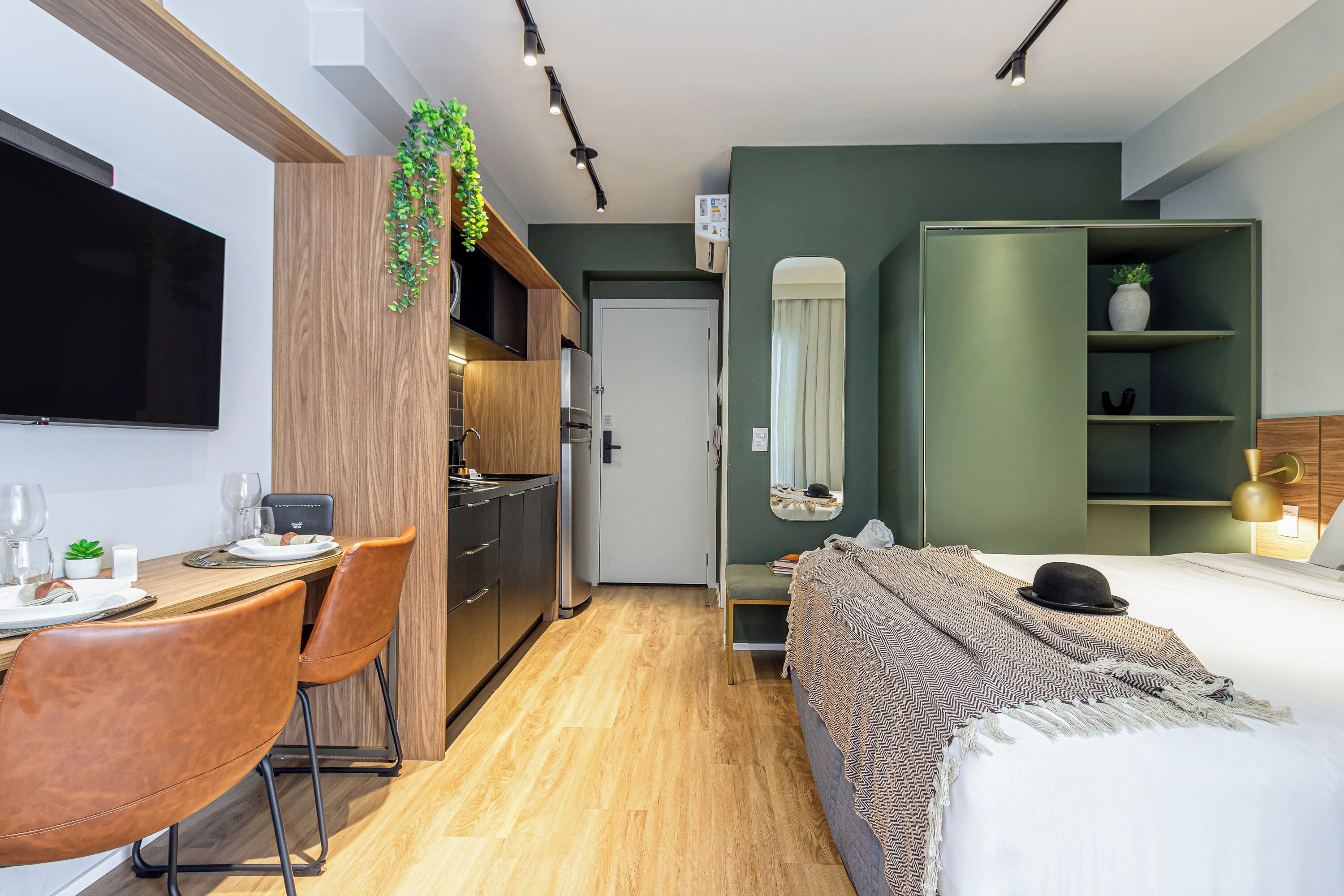 A compact hotel room with a kitchenette, a dining area, a bed with a decorative headboard, and a mirror above the bed.