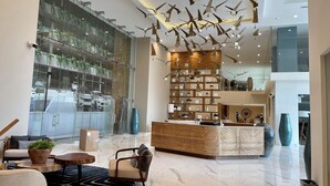 A modern hotel lobby with a central reception desk, a seating area with a sofa and armchairs, a large glass wall with a view of parked cars, and a ceiling with hanging light fixtures. - Luxury  2 Bedrooms Apartment at an exclusive tower by the beach in Zona Dorada 
 (Mazatlán)
