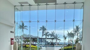 A modern lobby with a glass wall offering a view of a pool and palm trees. - Luxury  2 Bedrooms Apartment at an exclusive tower by the beach in Zona Dorada 
 (Mazatlán)