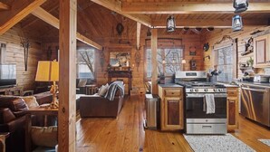 Een interieur van een chalet met een woonkamer, keuken en eethoek, met houten wanden en vloeren, een open haard en een groot raam.