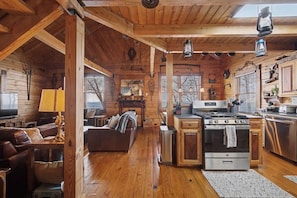 A cabin interior with a living room, kitchen, and dining area, featuring wooden walls and floors, a fireplace, and a large window. - Owl’s Hollow - Stunning Brow View (Mentone)