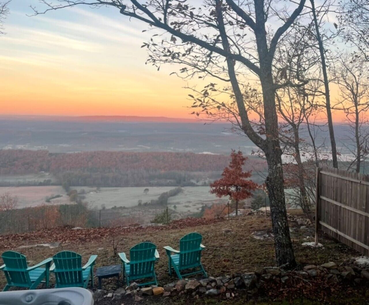 A view of a rural landscape with a sunset, a wooden fence, and green Adirondack chairs.