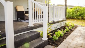 Eine überdachte Veranda mit weißem Geländer, einem Korbstuhl und einem kleinen Gartenbereich mit Pflanzen und Mulch.