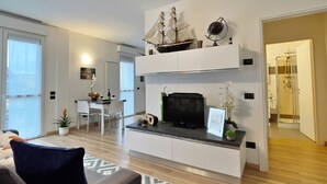 A modern living room with a sofa, a coffee table, a TV, and a ship model on the wall. There is a dining area with chairs and a table, and a bathroom with a shower. -  Rentopolis Apartments - Palazzo Candiani (Milan)