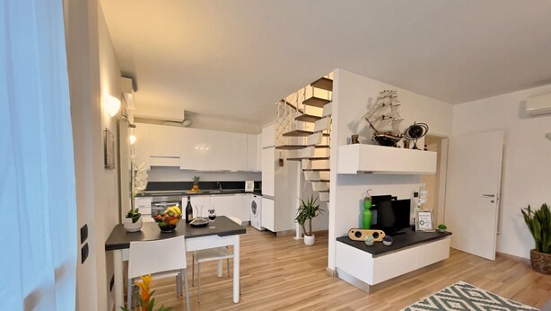 A modern kitchen with white cabinets, a dining area with a white table and chairs, and a living area with a patterned rug and a ship model on a shelf. -  Rentopolis Apartments - Palazzo Candiani (Milan)