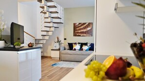 A modern living room with a staircase, a sofa with colorful cushions, a dining area with fruit, and a flat-screen TV. -  Rentopolis Apartments - Palazzo Candiani (Milan)