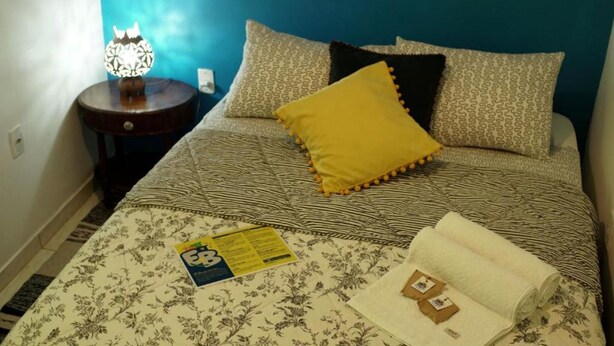 A neatly made bed with patterned bedding, a yellow pillow, and a small table with a lamp beside it. - Double Room with Shared Bathroom (Leme Rio de Janeiro)
