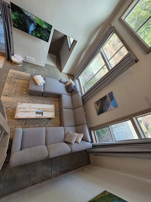 Een moderne woonkamer met een modulaire bank, een flatscreen televisie aan de muur en grote ramen die voor natuurlijk licht zorgen.