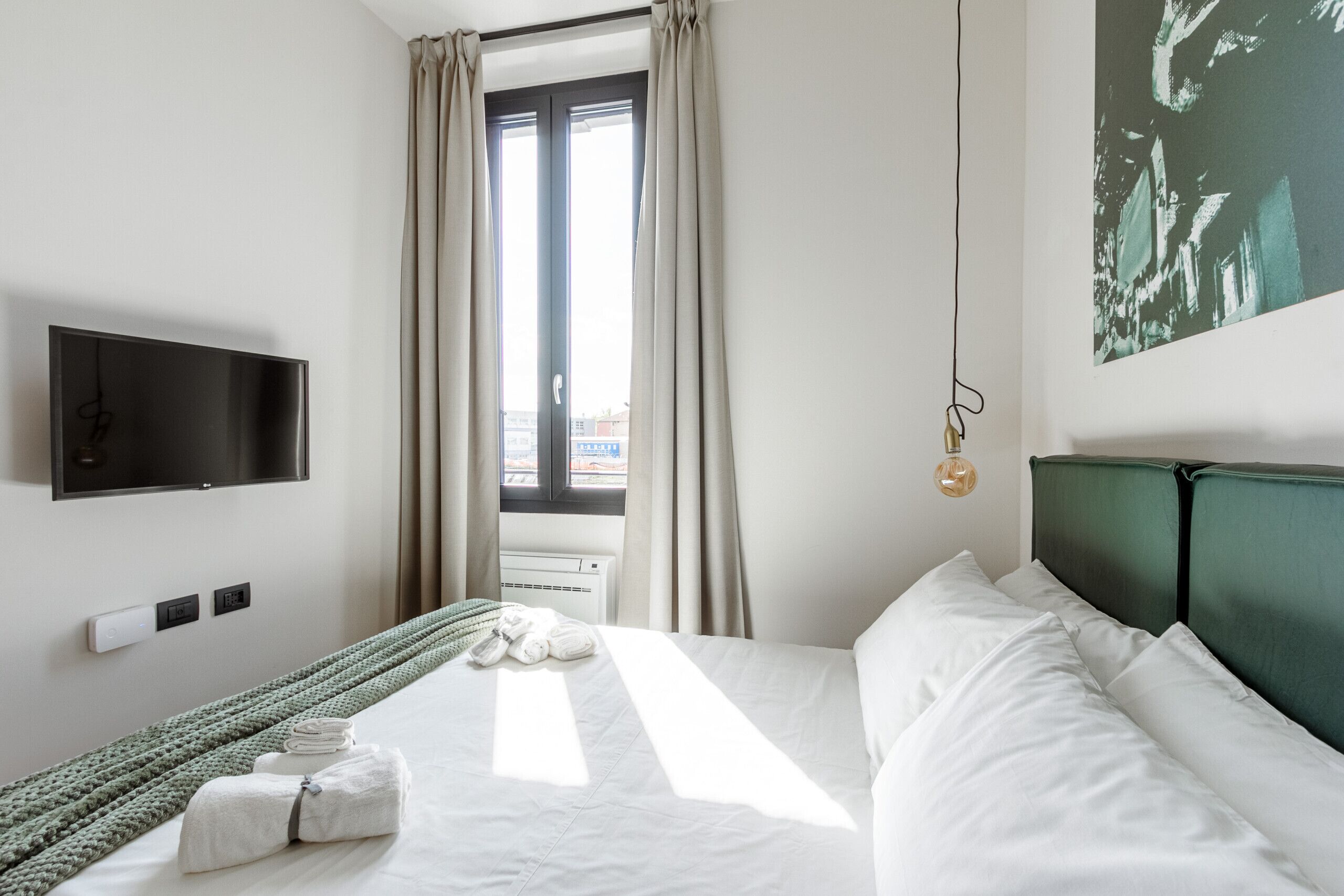 A hotel room with a bed, a flat-screen TV mounted on the wall, a window with curtains, and a painting on the wall.
