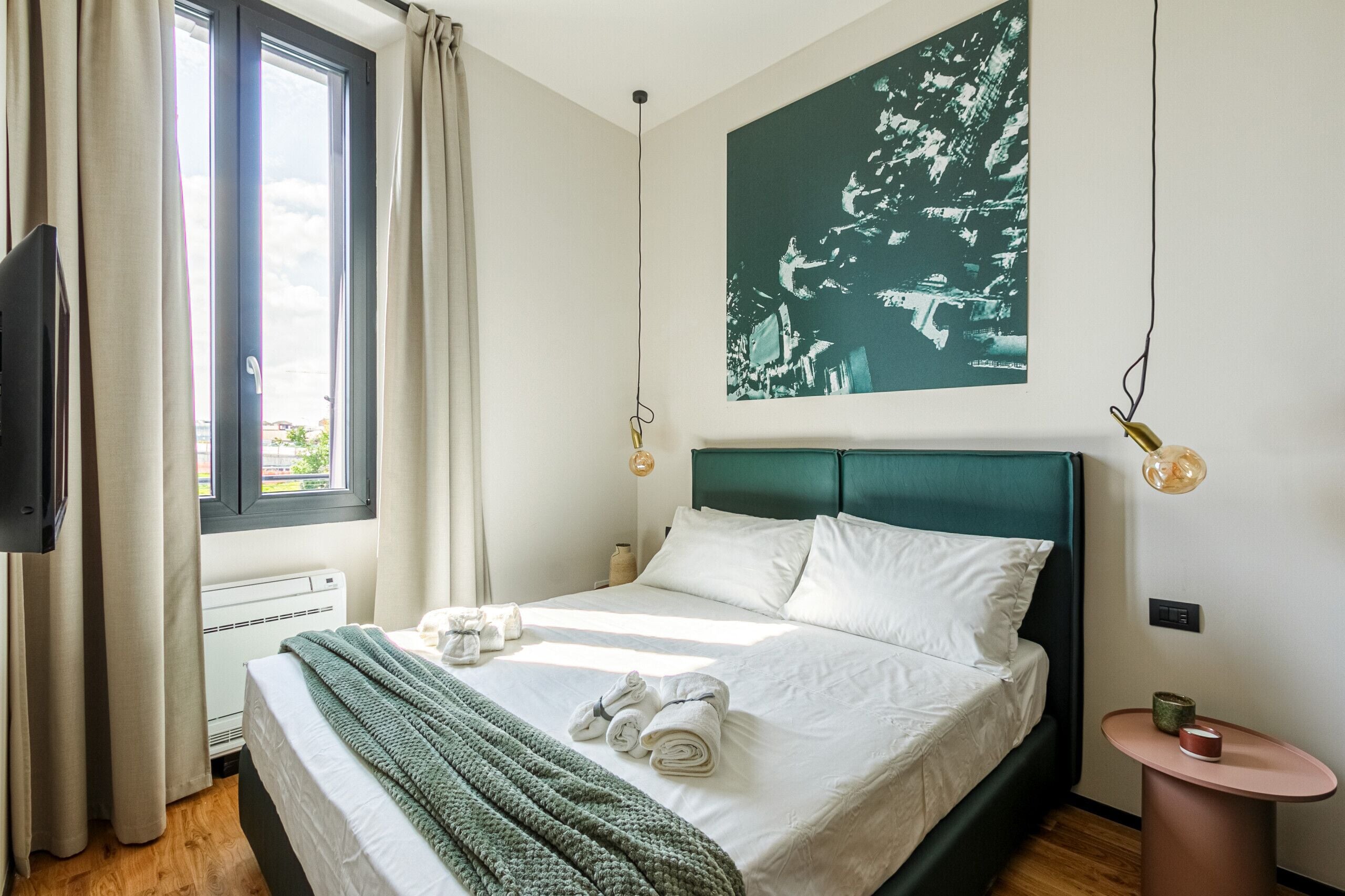 A neatly made bed with white linens, a green throw blanket, and rolled towels. A large abstract painting hangs above the bed.